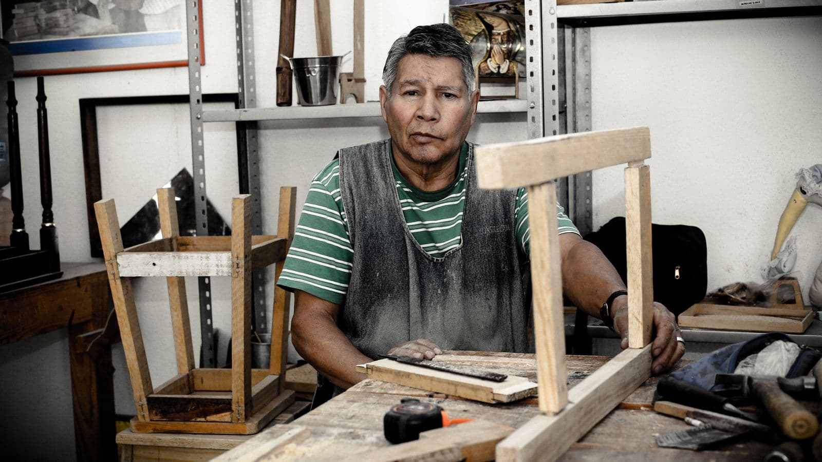 Un menuisier en tablier usé assemble un cadre en bois sur son établi, entouré d'outils et de tabourets en cours de fabrication