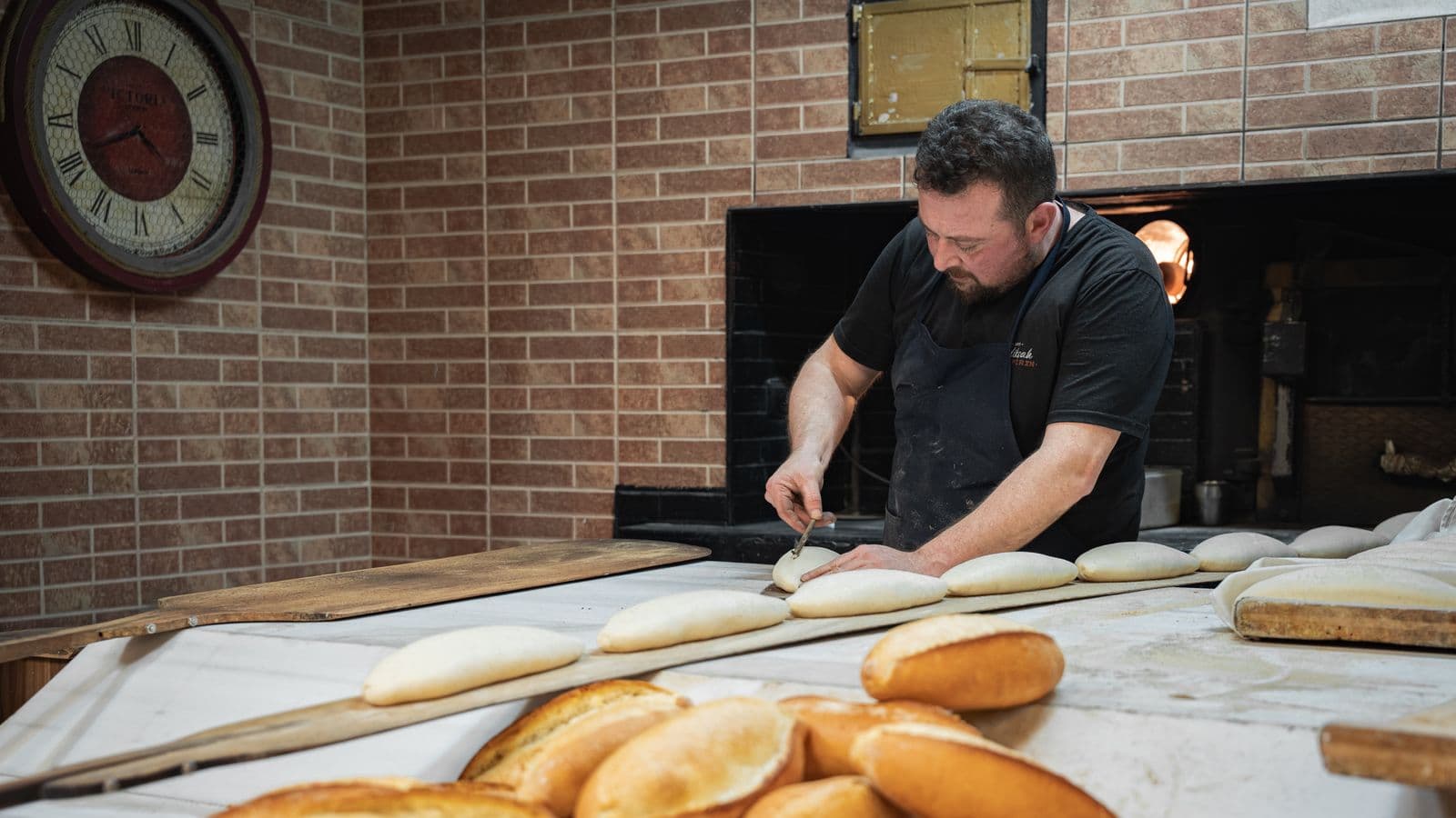Un boulanger en tablier façonne des pâtons sur une planche de bois devant son four, des pains dorés au premier plan